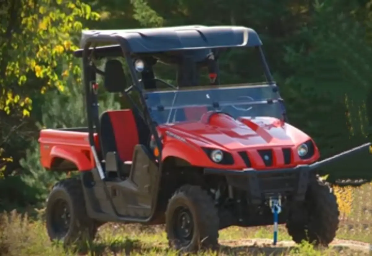 A red Yamaha Rhino 700 is parked on a grassy field. In the background there are trees and bushes.