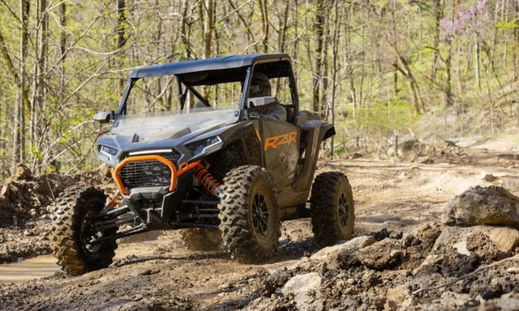 A man is riding a UTV on a muddy trail in the woods. He is going to a garage for weight comparison.