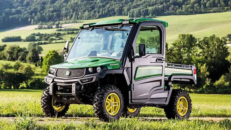 a John Deere Gator UTV is parked on a grassy field for weight comparison. The grass is green and lush, and there are trees and green field in the background.