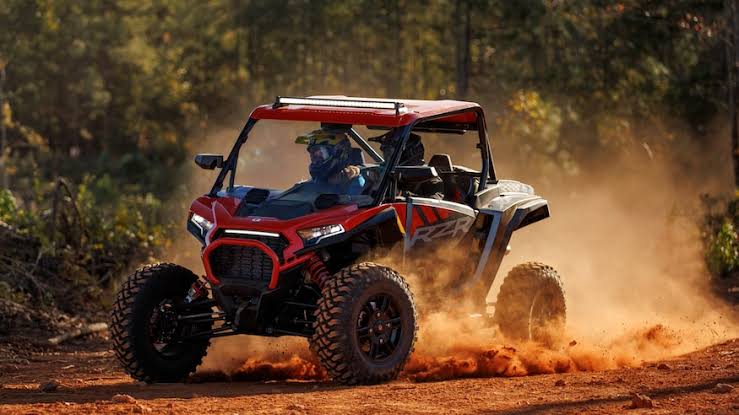 A person is riding a red UTV on a dirt road in a forest. The dirt road is winding and dusty, and there are trees on both sides.