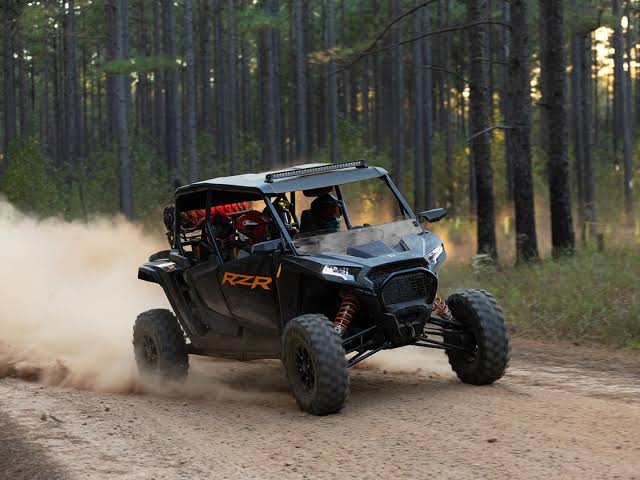 A man is riding a black Polaris RZR UTV on a dirt road in the forest. The road is dusty and in the background there are trees. The man is going to a UTV garage for UTV weight comparison.