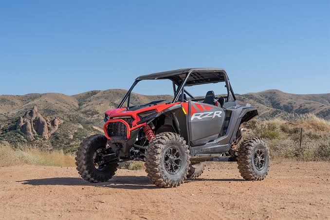 A red and black Polaris RZR UTV is parked on a dirt road in the desert for weight comparison. there are mountains in the background.
