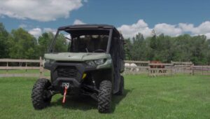 A Can-am defender pro XT HD10 is parked in a green field close to a farm. In the background there are trees and beautiful blue sky.