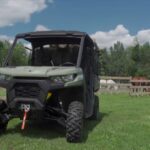 A Can-Am defender HD10 is parked in a green field in front of a farm. Its a beautiful sunny day. In the background there are trees and beautiful blue sky.