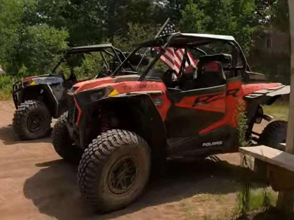 Two Polaris UTV parked on a dirt surface. In the background there are trees and bushes.