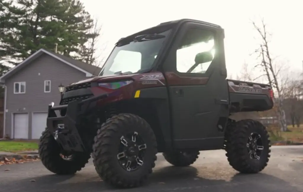 A Polaris Ranger UTV is parked on a road. In the background there is a house and trees.