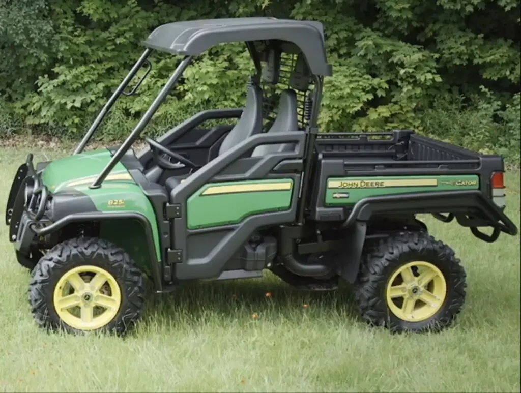 A John Deere gator is parked on a grassy field. In the background there are trees and bushes.