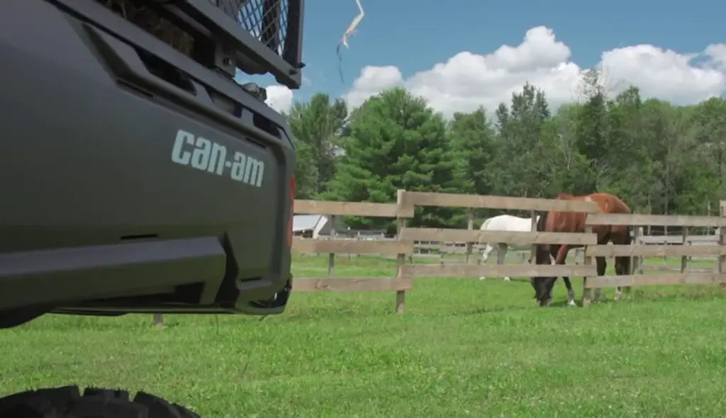 A Can-am defender HD10 is parked next to a green field with fence. In the green field two horses are eating grass. In the background there are trees and beautiful blue sky.