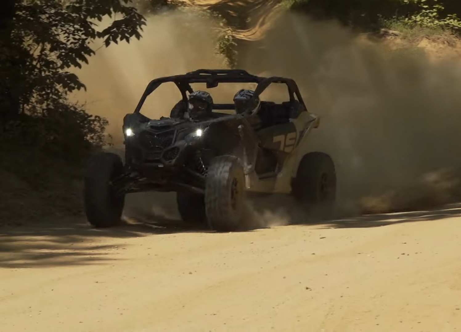 Two people are riding on the Can-am Maverick X3 through a dusty trail.