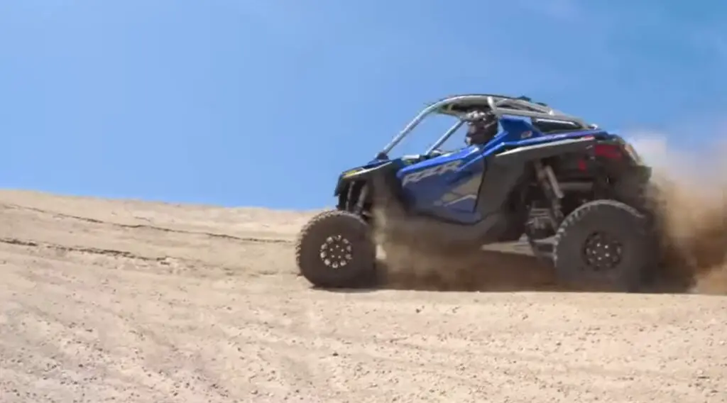 A Polaris RZR is being driven through a sandy surface.