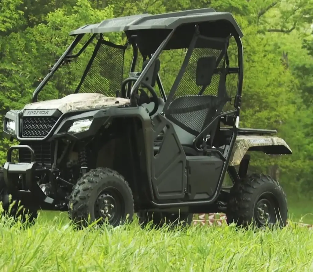 A Honda Pioneer 500 is parked in a grassy field next to a green jungle.