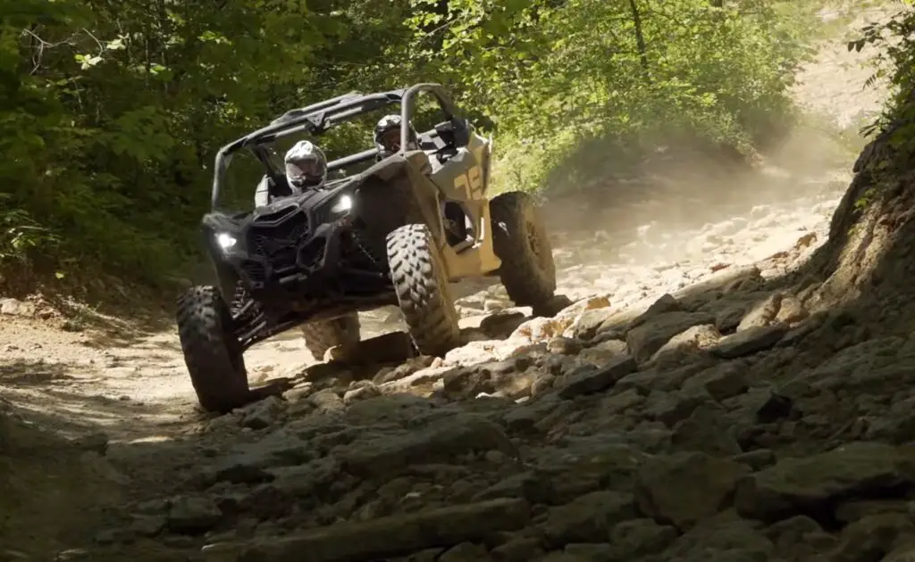 A Can-am Maverick X3 is being driven through a gravel road.