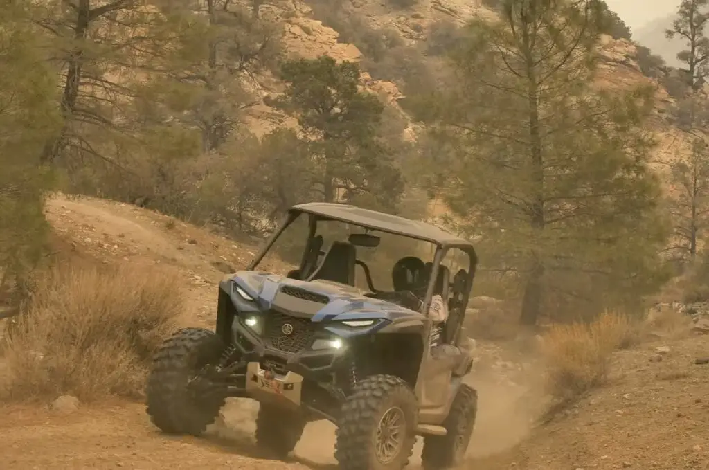 A Yamaha Wolverine RMAX2 is being driven through a dirt and dusty road. In the background there are trees and mountains.