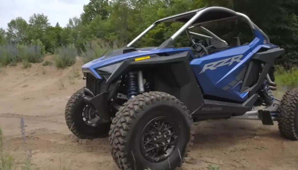 A Polaris RZR is parked on a sandy surface. In the background there are trees and bushes.