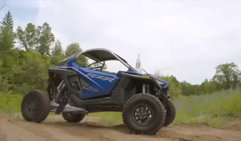 A Polaris RZR UTV is parked on a dirt surface in front of a green field.