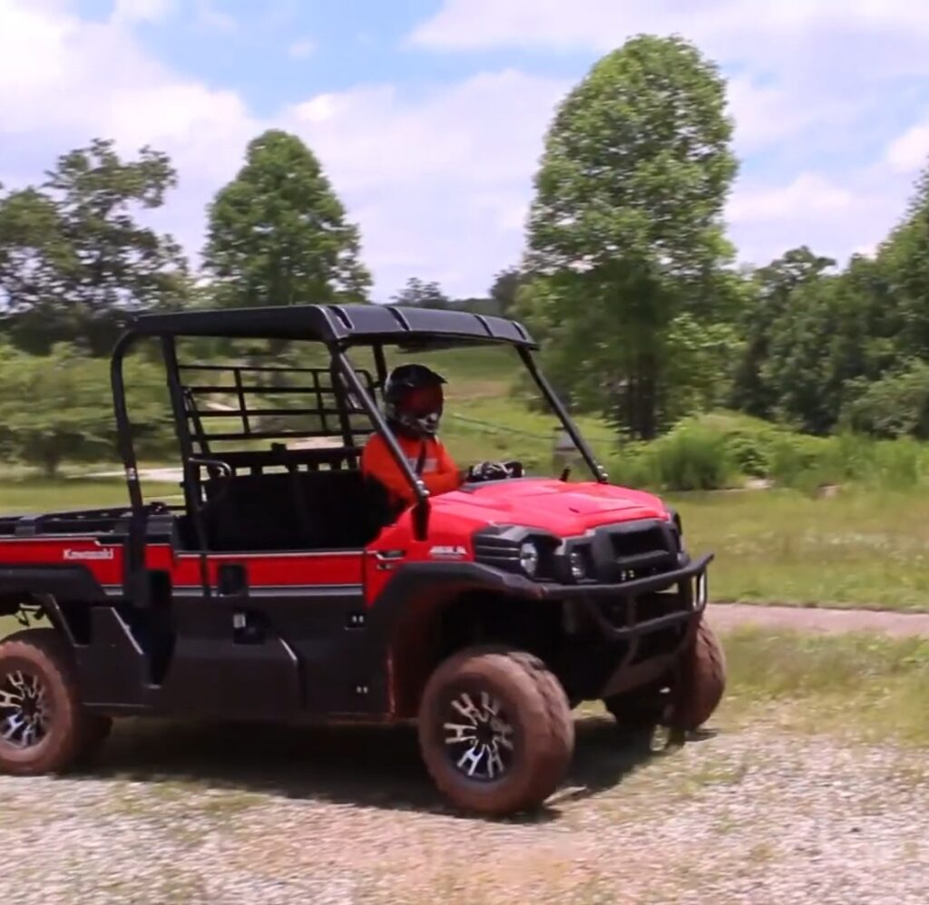 A Kawasaki Mule Pro-FX EPS LE is being driven through a dirt road. In the background there are trees and beautiful sky.