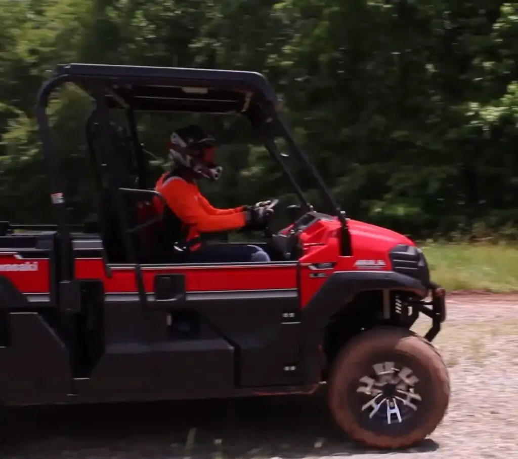 A Kawasaki Mule Pro-FX EPS LE is being driven through a dirt road. In the background there are trees and bushes.