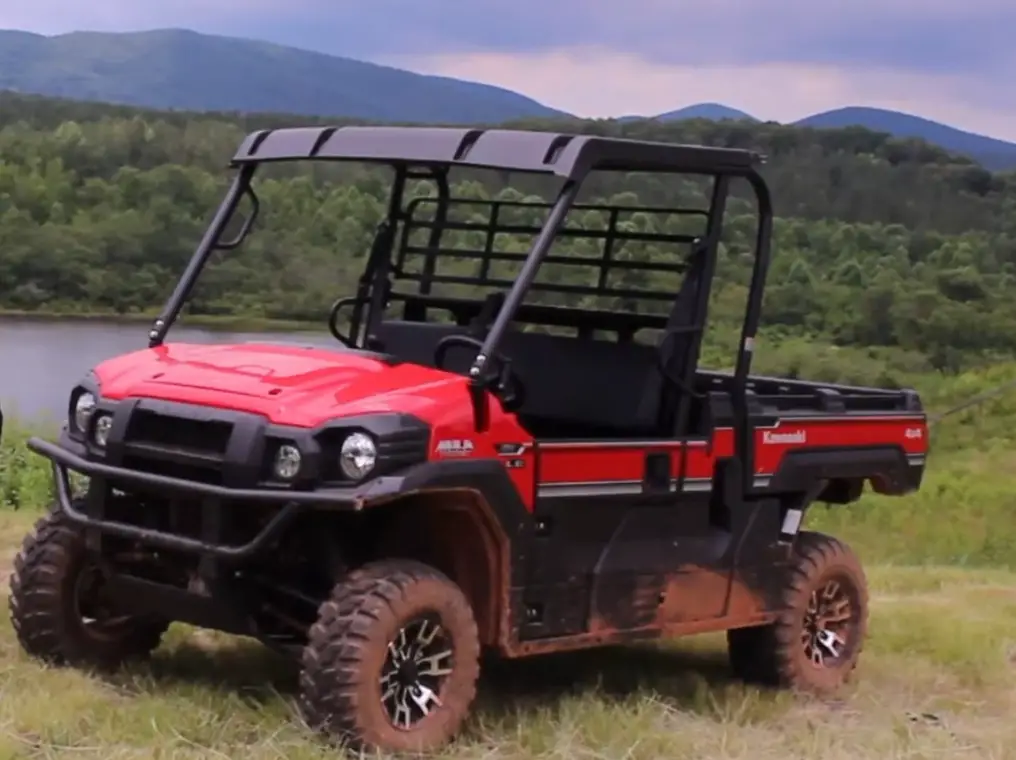 A Kawasaki Mule Pro-FX EPS LE is parked in front of a lake. In the background there is a green forest and mountains.