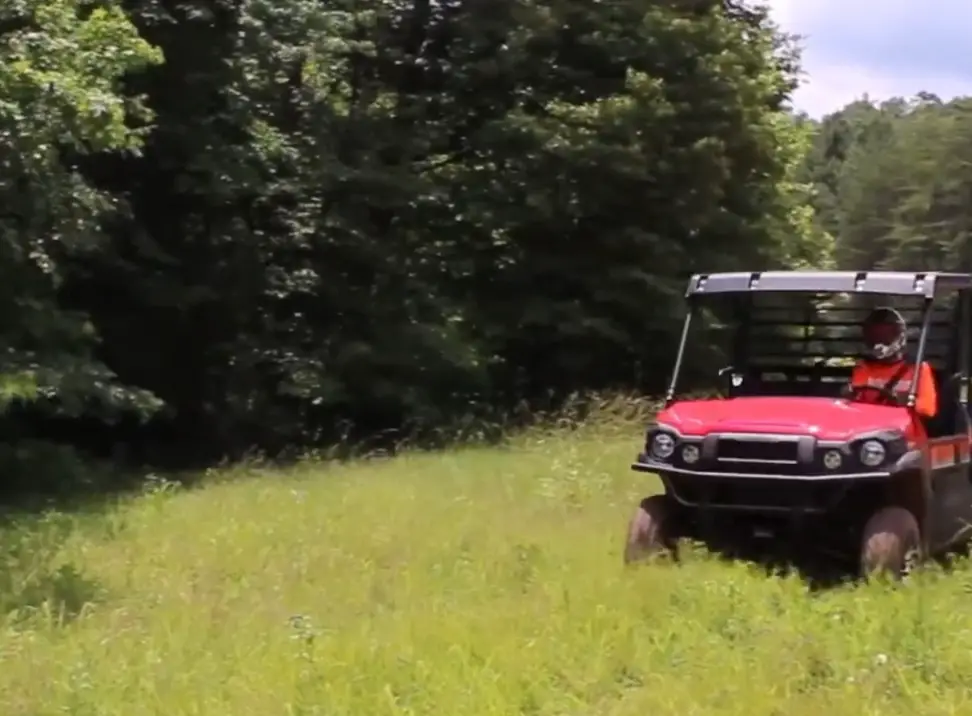 A Kawasaki Mule Pro-FX EPS LE is being driven through a grass field. In the background there are trees and bushes.