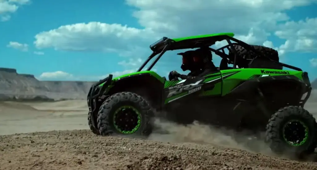 A Kawasaki Teryx KRX 1000 is being driven through a dusty road in desert.