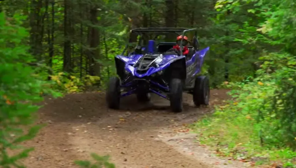 A blue Yamaha YXZ1000R is being driven through a dirt road in a jungle.