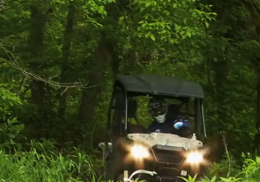 A Honda Pioneer 500 with lights on is being driven through a jungle terrain.