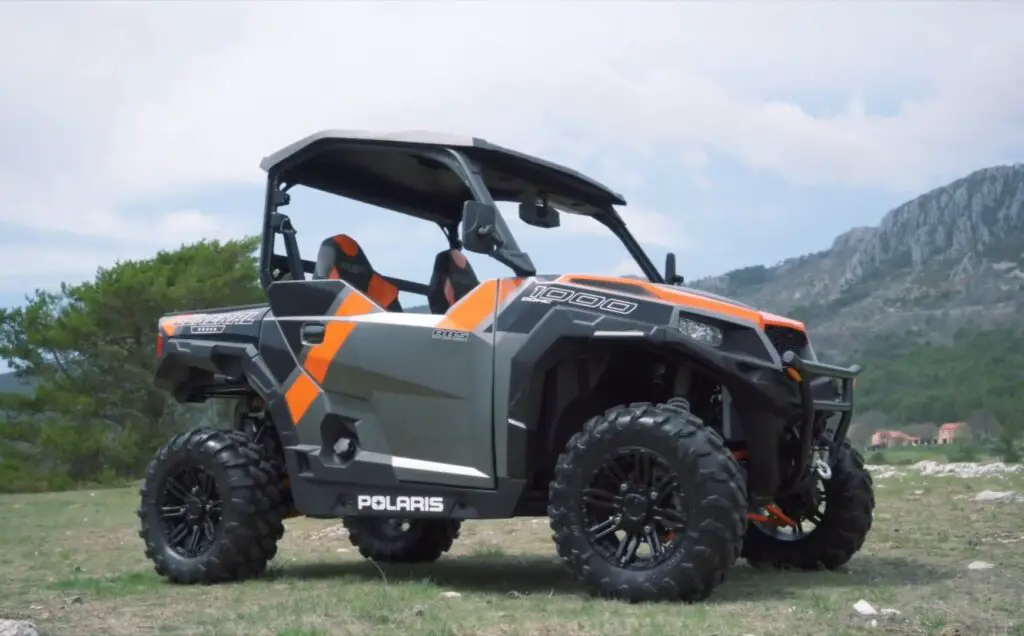 Polaris General 1000 EPS is parked on a grassy field. In the background there are trees, mountains and white sky with clouds.