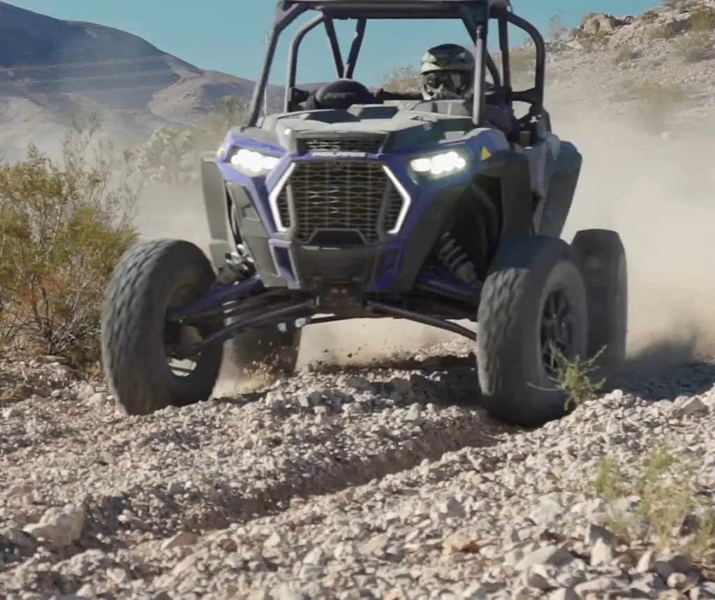 A Polaris RZR XP Turbo S is being driven through a gravel road. In the background there are mountains and beautiful blue sky.