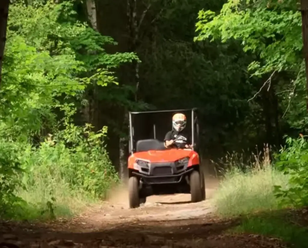 A Polaris Ranger 500 is being driven through a dirt road. There are trees and bushes on the both side of the road.